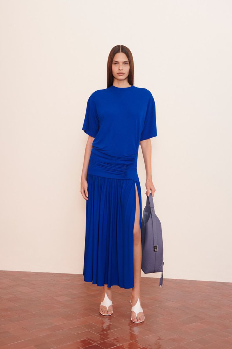 Woman in blue t-shirt dress with side slit stands indoors holding blue-grey handbag; wears white sandals, neutral backdrop, and tile floor.