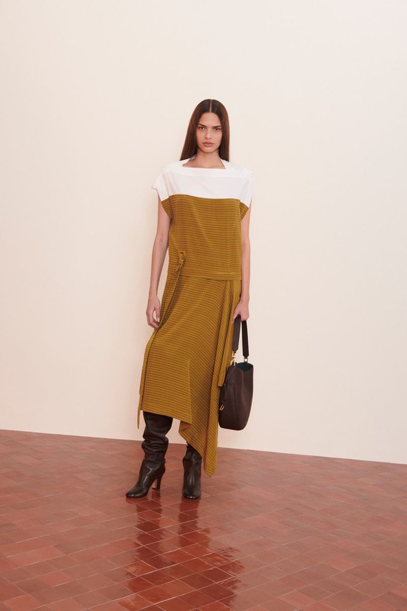 Full-length shot of a woman in a two-tone white and mustard dress with long sleeves, black heeled boots, and a brown purse.