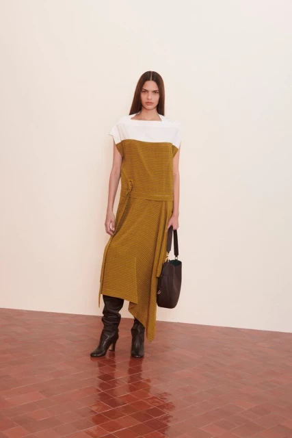 Full-length shot of a woman in a two-tone white and mustard dress with long sleeves, black heeled boots, and a brown purse.