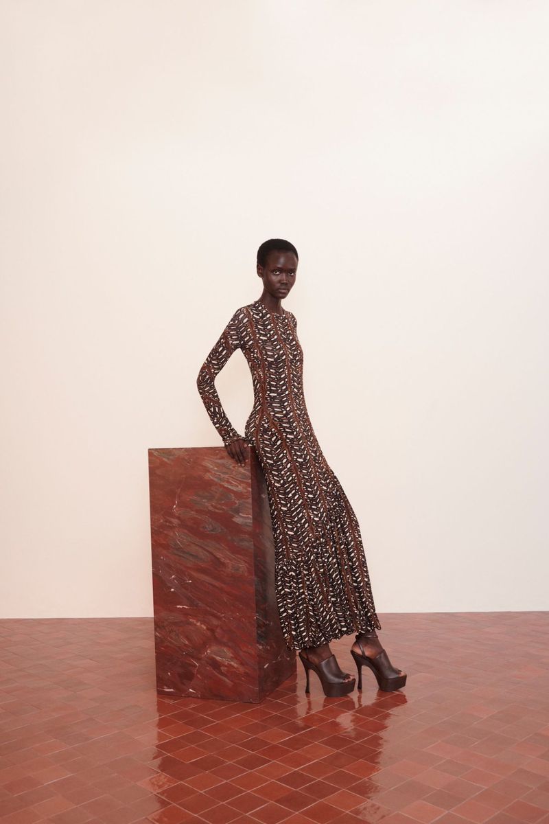 A Black woman in a printed maxi dress and black platform heels poses in a studio setting, leaning on a geometric block.