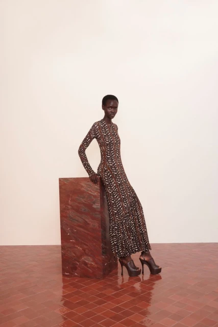 A Black woman in a printed maxi dress and black platform heels poses in a studio setting, leaning on a geometric block.