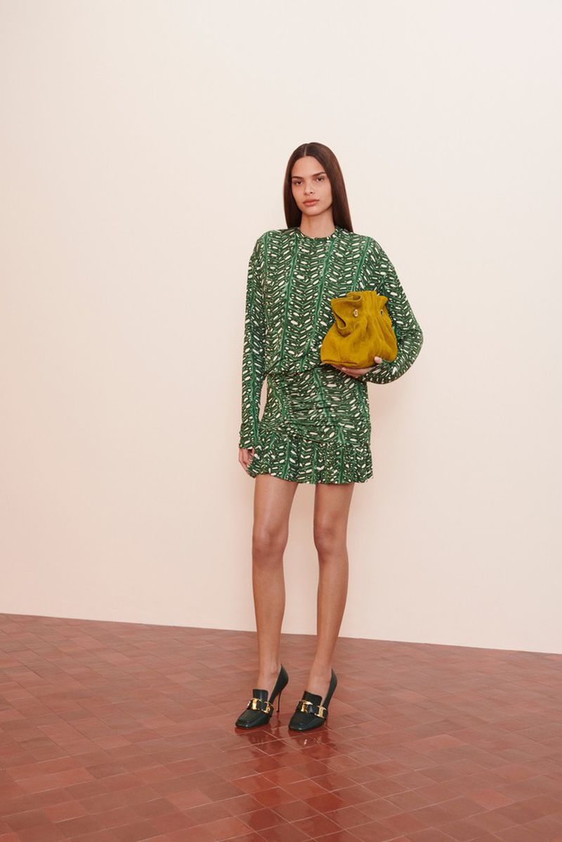 A woman in a green patterned dress holds a yellow purse, standing in a studio with a neutral background and red brick floor.