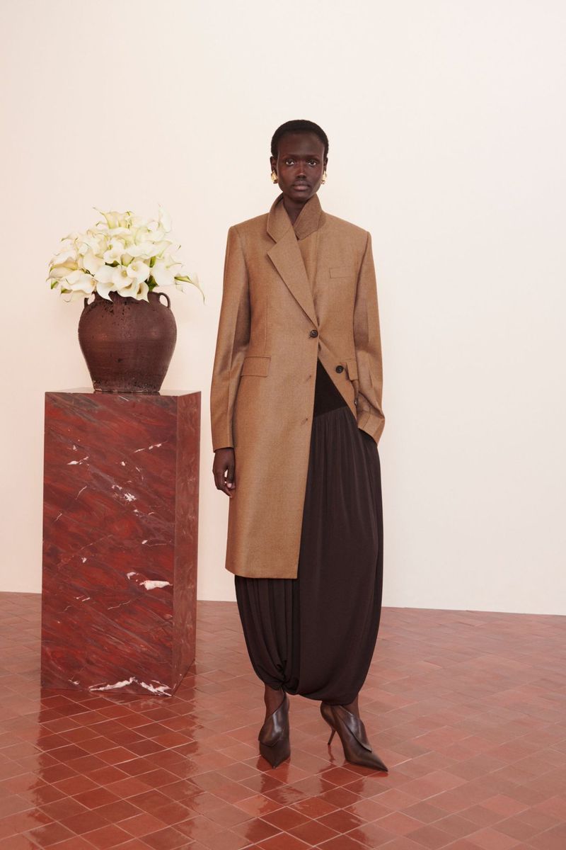 A full-length shot of a Black female model wearing a tan coat, black harem pants and heels, standing indoors near a floral arrangement.