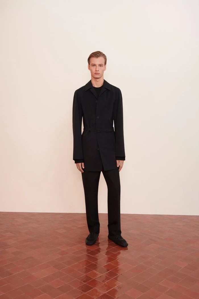 Model standing on a tiled floor against a plain wall wearing a black jacket and matching trousers.