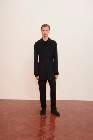 Model standing on a tiled floor against a plain wall wearing a black jacket and matching trousers.