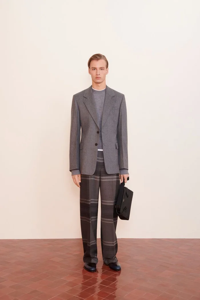 Full-length portrait of a male model wearing a grey blazer, patterned trousers, and holding a black handbag.