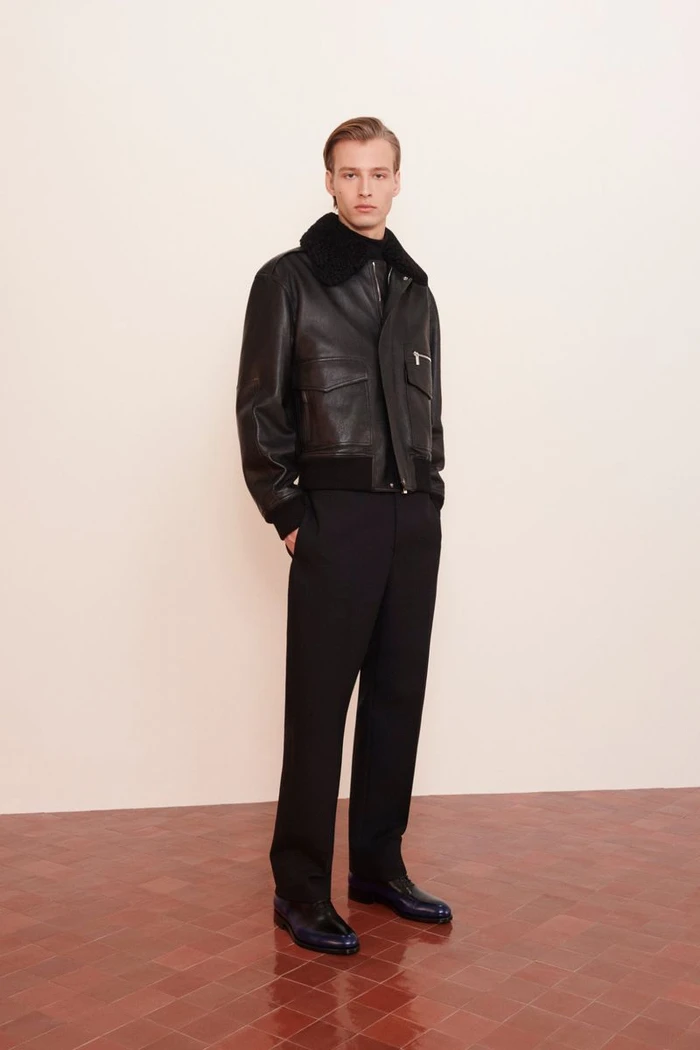 Model wearing a black leather jacket with a shearling collar and black trousers against a neutral background