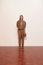 Model wearing a brown belted coat and trousers holding a brown leather handbag in a studio