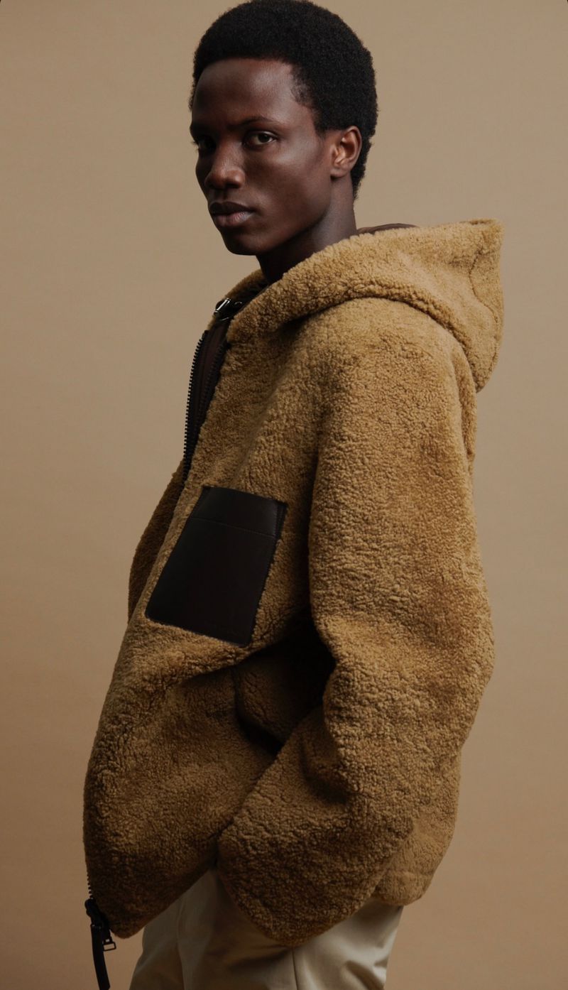 Man in a tan hooded sherpa jacket posing in a studio. The jacket has a black patch on the front and is zipped up.