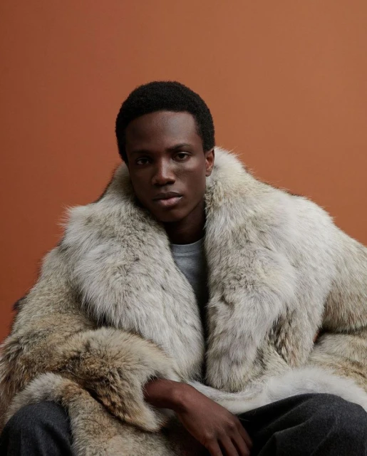 An adult Black man wearing a large fur coat poses in a studio with a bronze background, showcasing fashion and style.