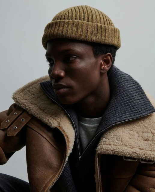 Portrait of a stylish adult Black man in a tan beanie and shearling-lined jacket, against a light background, looking to the side.