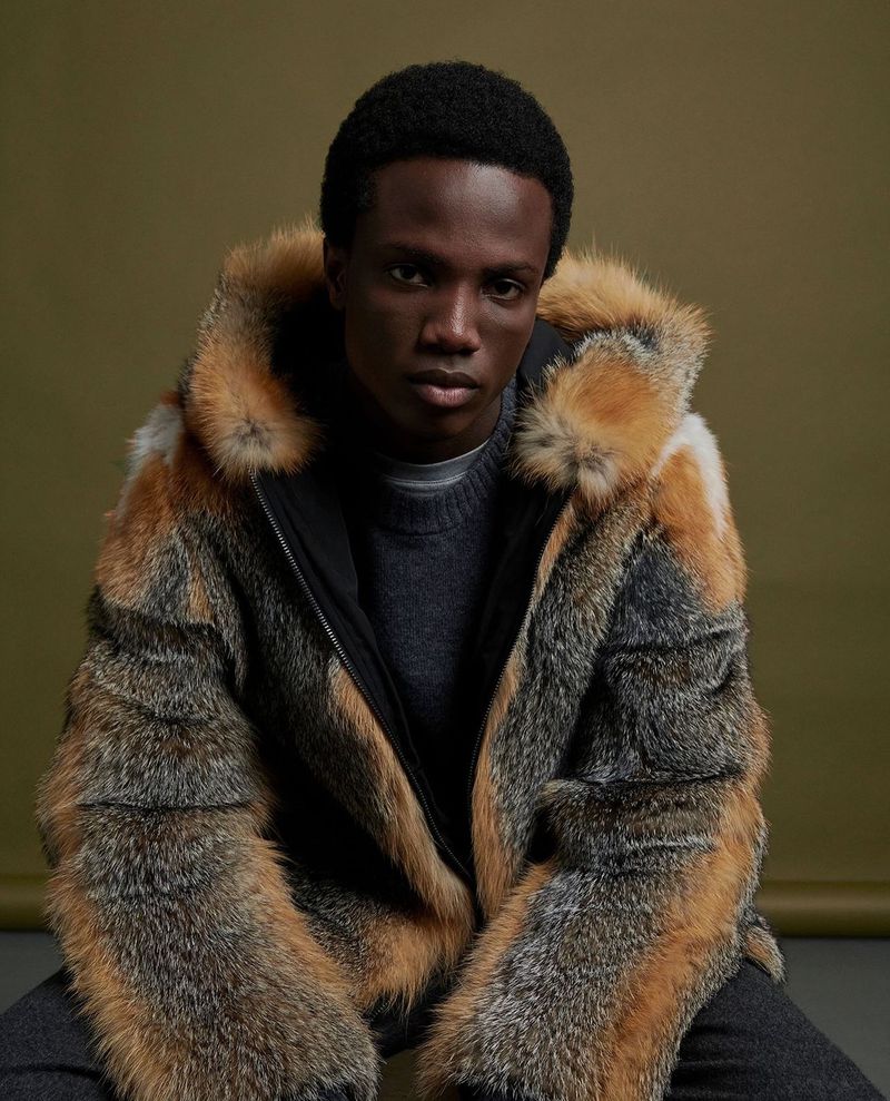 Adult male poses indoors wearing a fur coat and grey sweater against an olive-green backdrop in a fashion portrait.