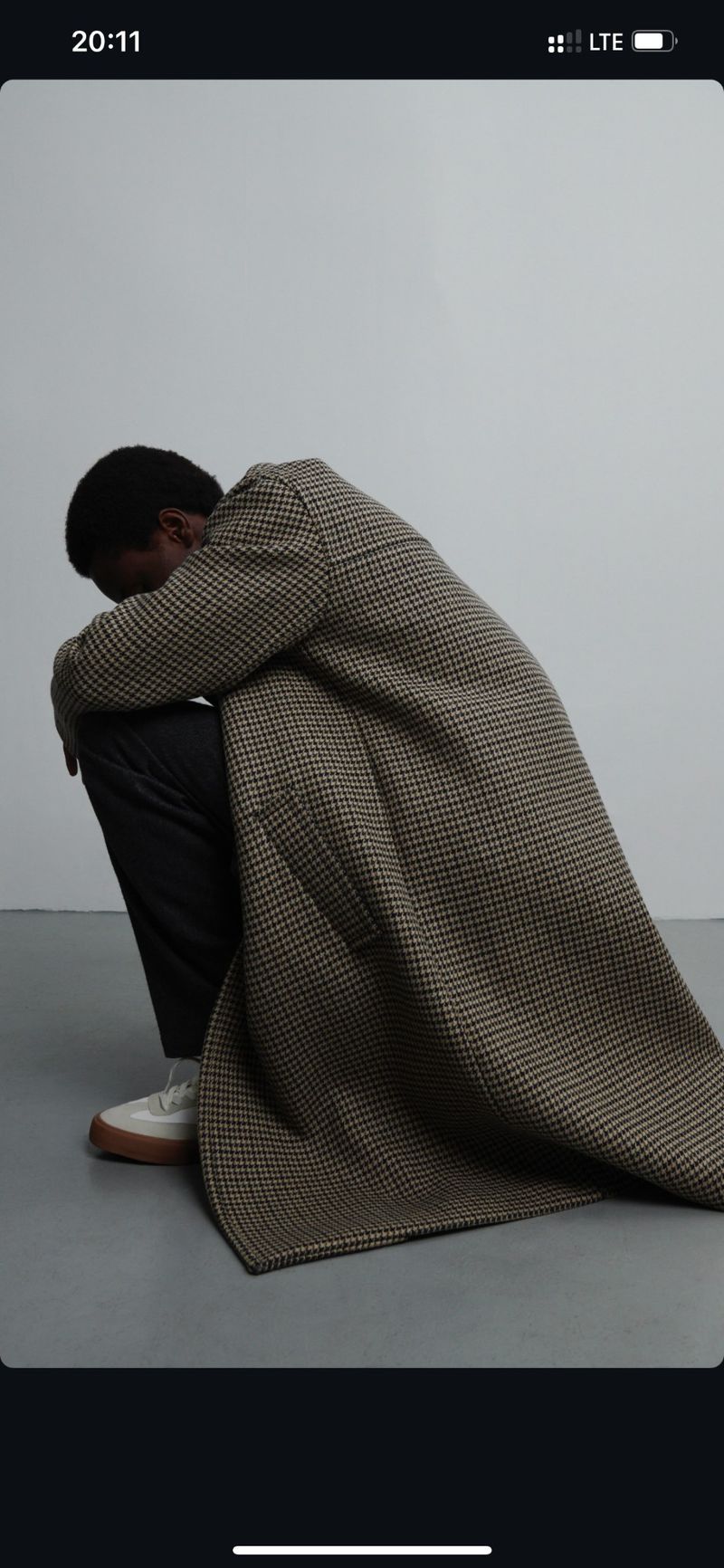 An African-American man sits, head down, wearing a patterned coat, dark pants, and light-colored shoes against a neutral background.