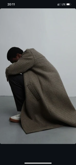 An African-American man sits, head down, wearing a patterned coat, dark pants, and light-colored shoes against a neutral background.
