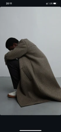 An African-American man sits, head down, wearing a patterned coat, dark pants, and light-colored shoes against a neutral background.