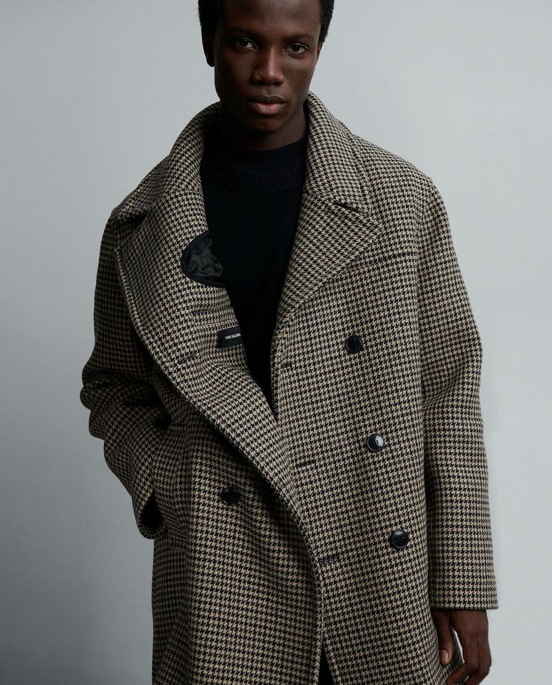 A dark-skinned adult male poses indoors, wearing a patterned overcoat and black top, in a medium shot with soft lighting against a grey background.