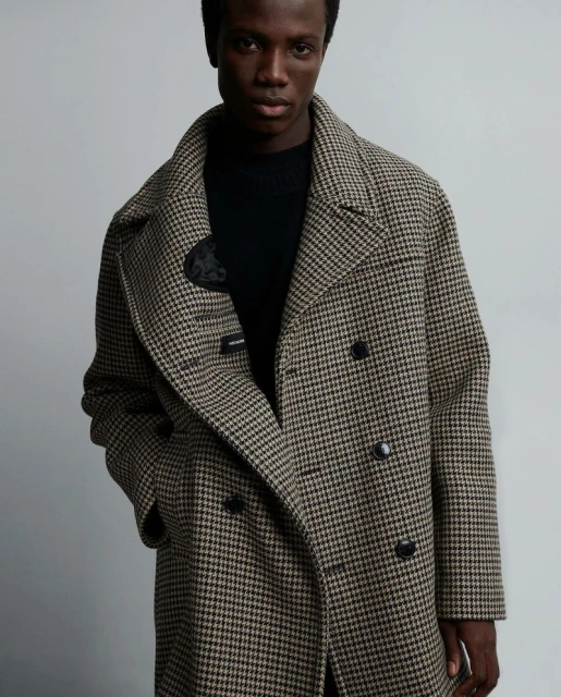 A dark-skinned adult male poses indoors, wearing a patterned overcoat and black top, in a medium shot with soft lighting against a grey background.
