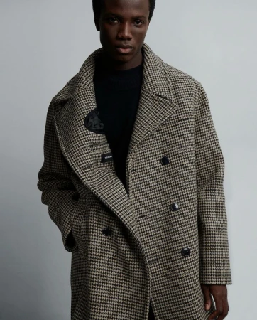 A dark-skinned adult male poses indoors, wearing a patterned overcoat and black top, in a medium shot with soft lighting against a grey background.