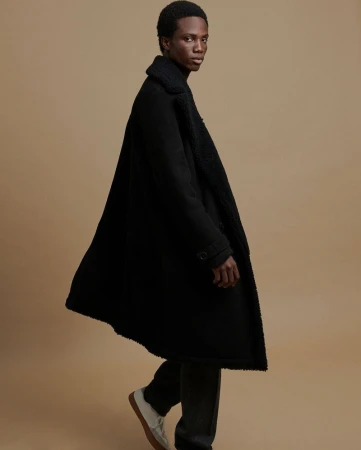 Portrait of a young adult Black male model wearing a full-length black wool coat with beige shoes, posing against a tan backdrop.