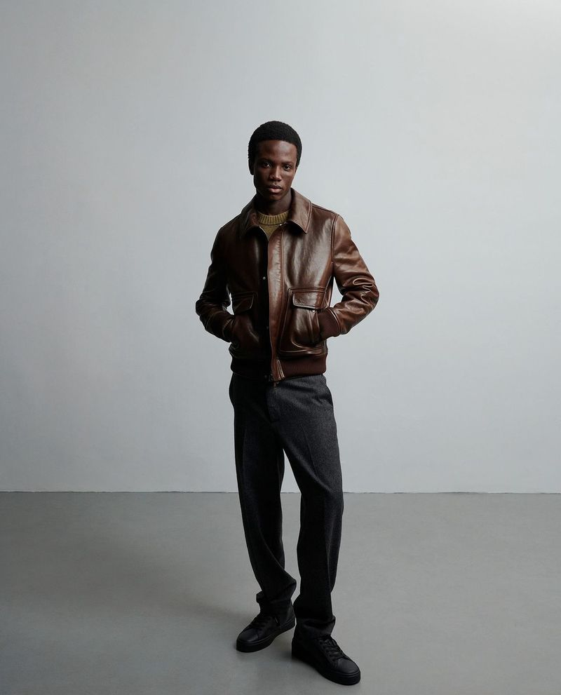 A male model of African descent poses in a brown leather bomber jacket, gray trousers, and black leather shoes against a gray background.