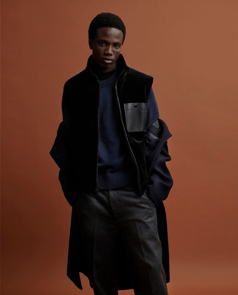 Stylish African-American man in layered dark blue and black vest and overcoat fashion in a studio shot against a brown background.