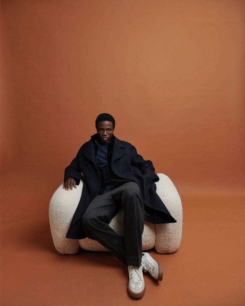 A Black man in a coat sits in a white chair against a brown backdrop, modelling streetwear and menswear fashion.