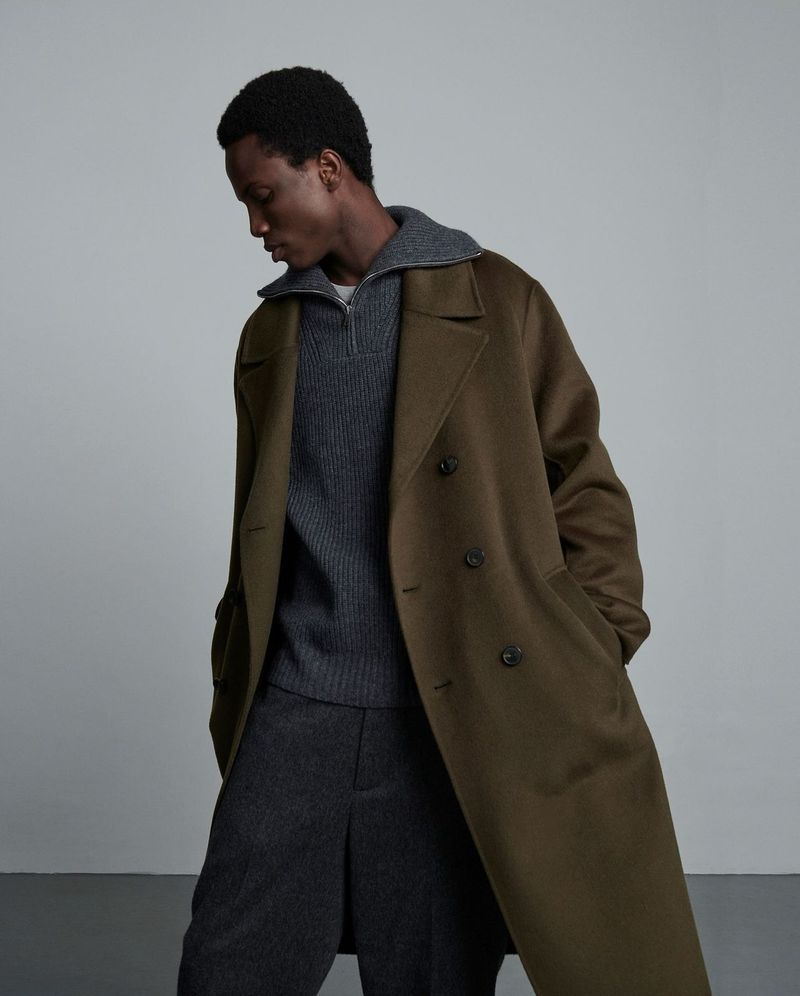 Mens fashion: A dark-skinned man in a grey sweater and olive overcoat poses in a studio setting with a minimalist background.