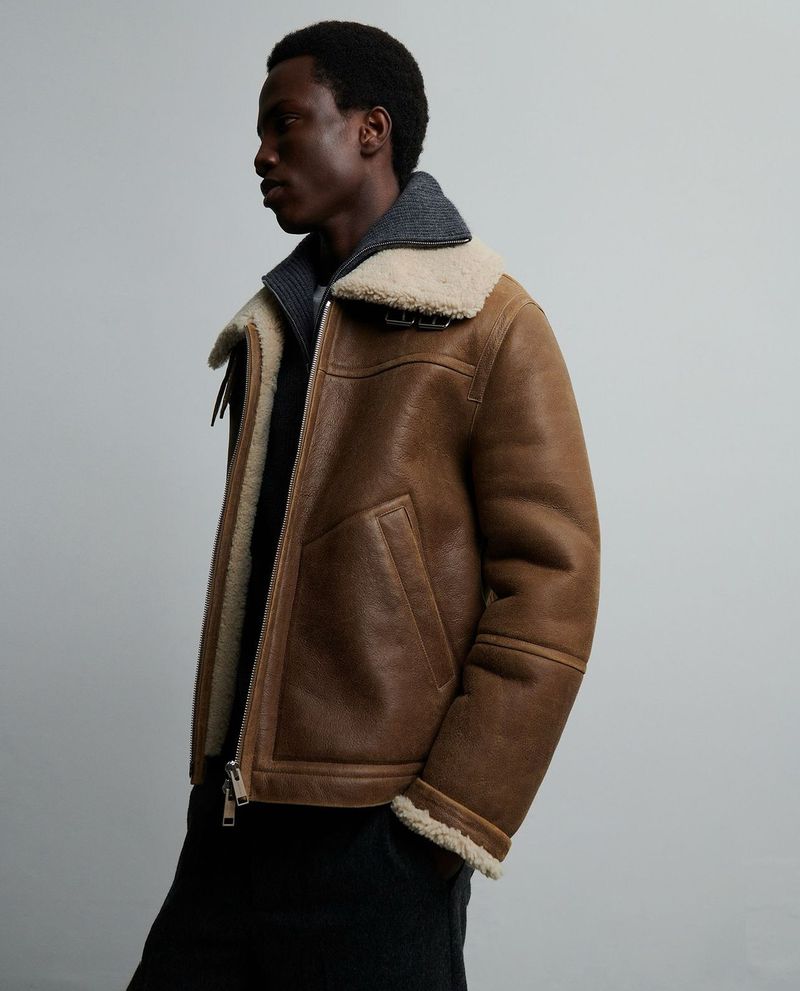Adult male wearing brown aviator jacket with cream shearling collar and grey wool top against a neutral grey backdrop in studio lighting.