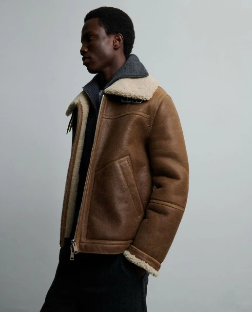 Adult male wearing brown aviator jacket with cream shearling collar and grey wool top against a neutral grey backdrop in studio lighting.