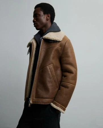 Adult male wearing brown aviator jacket with cream shearling collar and grey wool top against a neutral grey backdrop in studio lighting.