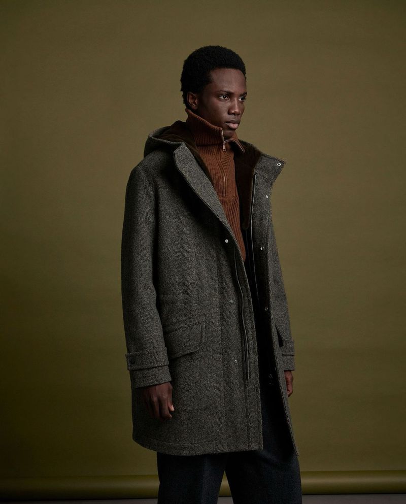A stylish, dark-skinned male model in a hooded grey tweed coat and brown ribbed turtleneck, is set against a muted olive green background.