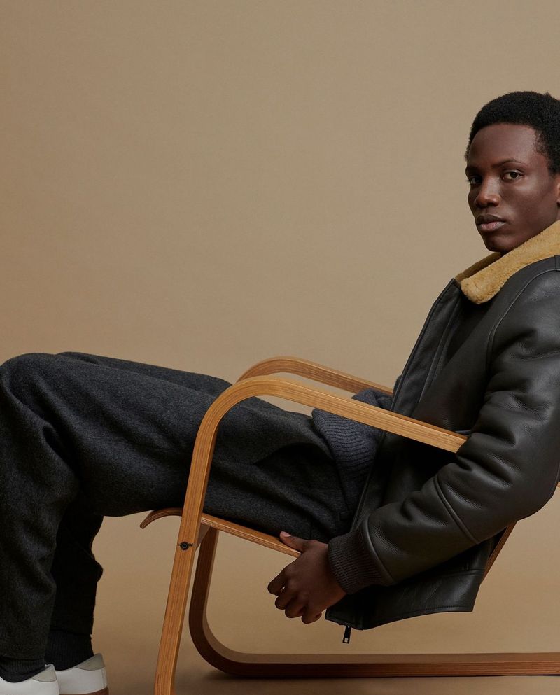 A Black man sits in a wooden chair wearing a leather jacket and dark pants in a studio with a tan background for a fashion shot.