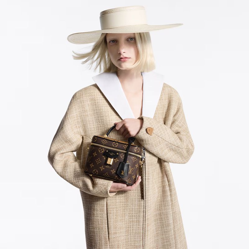 Fashion model in beige coat and wide-brimmed hat holds a Louis Vuitton vanity bag against a white background, studio lighting, close up.