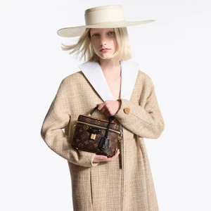 Model wearing a structured beige tweed coat and wide-brimmed cream hat holding a monogram handbag against white background