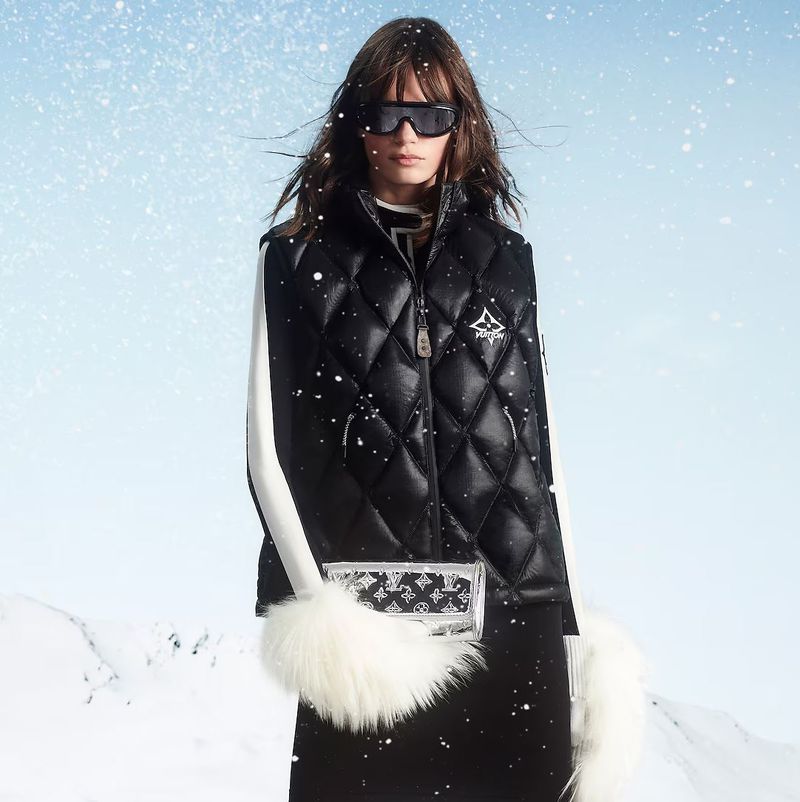 A brunette fashion model in a black vest poses in a snowy landscape. She wears black sunglasses, and a fur-trimmed Louis Vuitton bag.