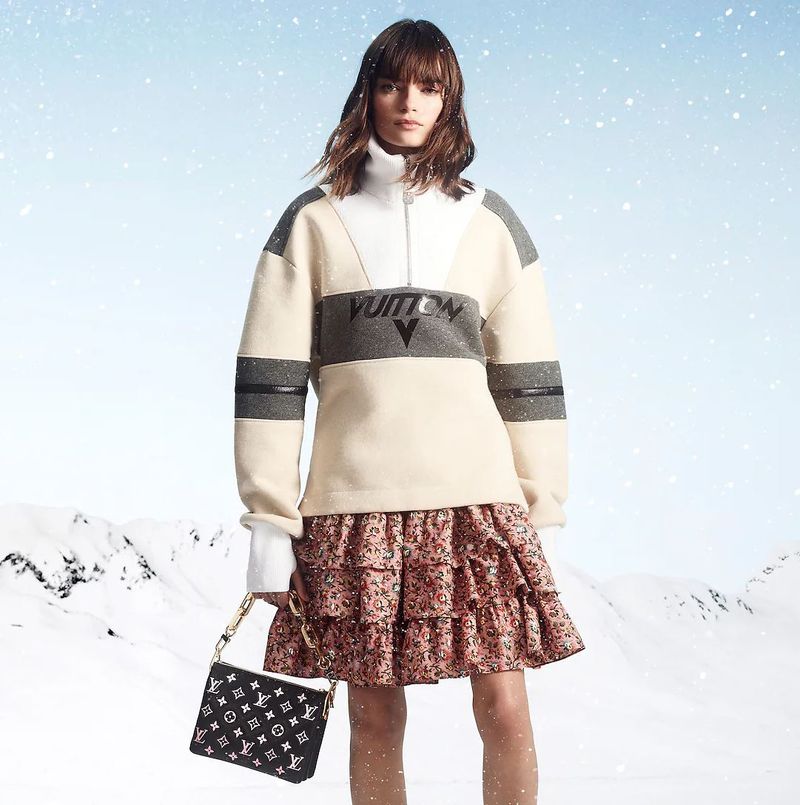 A woman poses in a cream-colored pullover and tiered floral skirt, carrying a Louis Vuitton handbag, set against a snowy mountain backdrop.