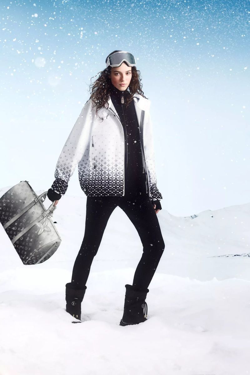 Woman in winter fashion on snowy landscape, wearing ski goggles, jacket, leggings, and boots, carrying luxury handbag, under a blue sky.