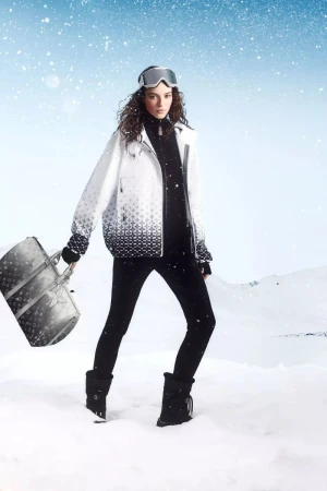 Woman in winter fashion on snowy landscape, wearing ski goggles, jacket, leggings, and boots, carrying luxury handbag, under a blue sky.