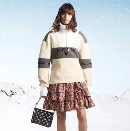 Model wearing a half-zip pullover and patterned skirt holding a black handbag against a snowy landscape background.