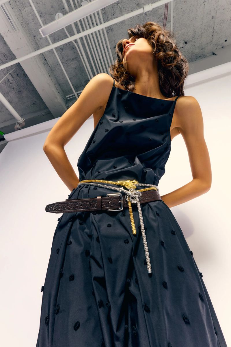 Fashionable woman posing in black sleeveless dress with brown, silver and gold belts in a photo studio with white walls.