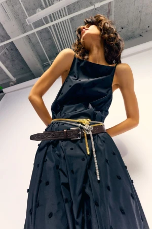 Fashionable woman posing in black sleeveless dress with brown, silver and gold belts in a photo studio with white walls.