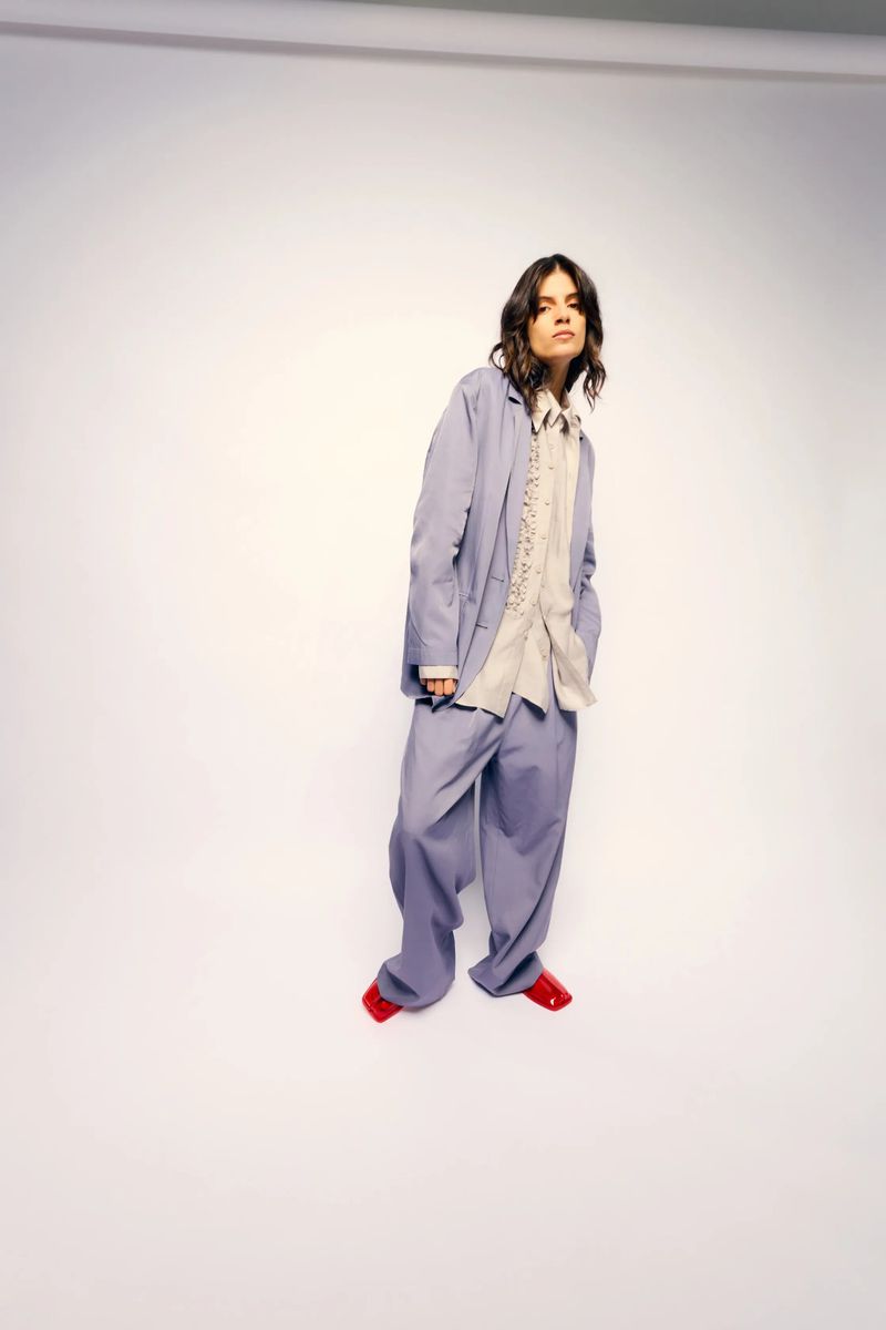 Adult woman in oversized lilac suit posing in studio with relaxed posture. She wears red flats and a cream-colored lace-trimmed top.