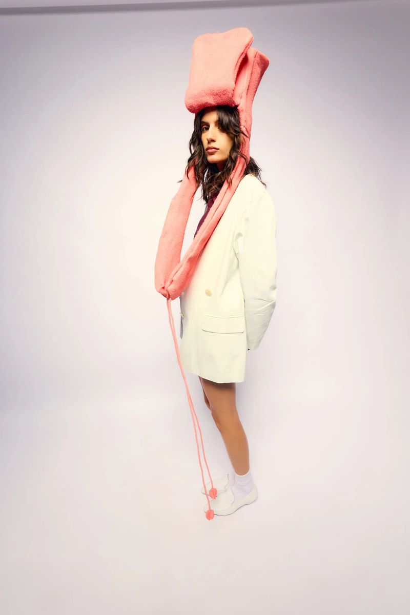 Woman in white blazer poses in front of a white background wearing a tall pink hat with long hanging strings and white sneakers.