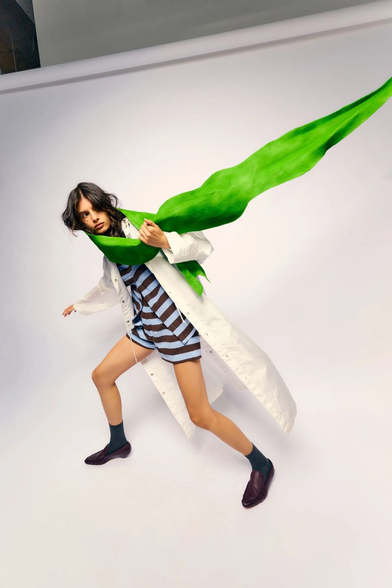 A woman strikes a dynamic pose on a white backdrop, adorned in a striped blue and brown outfit, accessorized with a white lab coat and a large green leaf.
