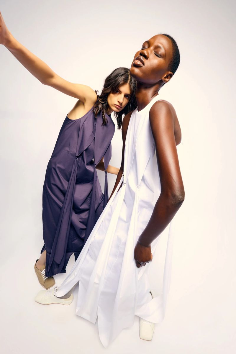 Two female models posing against a white background for Tibi Pre-Fall 2025, wearing sculptural dresses and sneakers.
