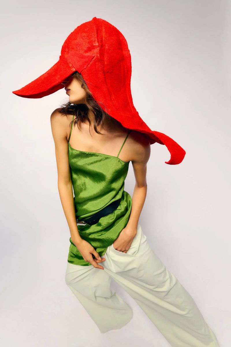 Model wearing a large red sculptural hat, green camisole top, and white trousers against a plain white studio backdrop.