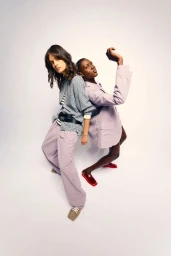 Two fashion models posing back-to-back against a plain white studio background wearing patterned shirts and trousers