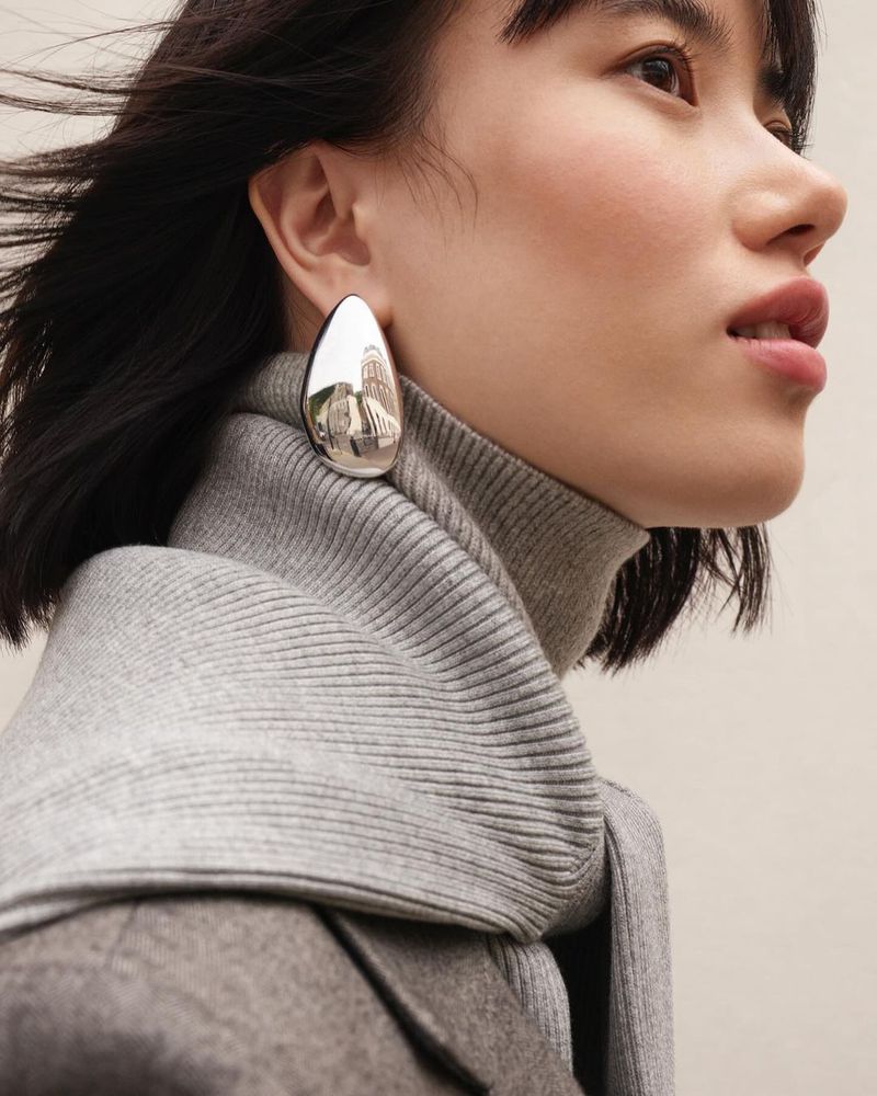 Asian woman in grey turtleneck sweater and wool coat wearing a silver teardrop earring, posing in a close-up studio portrait.