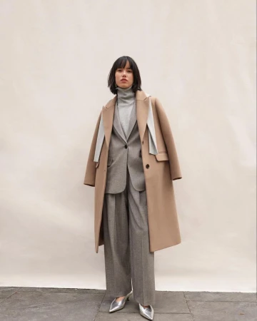 Full-length shot of a woman in a tan coat over a gray suit and turtleneck, standing against an off-white backdrop, complemented by silver heels.