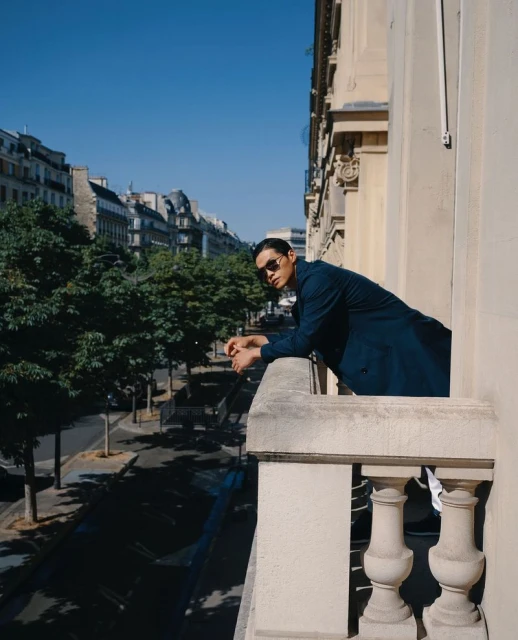 An adult male wearing a dark suit and sunglasses leans against a balcony railing overlooking a European street with trees and buildings.